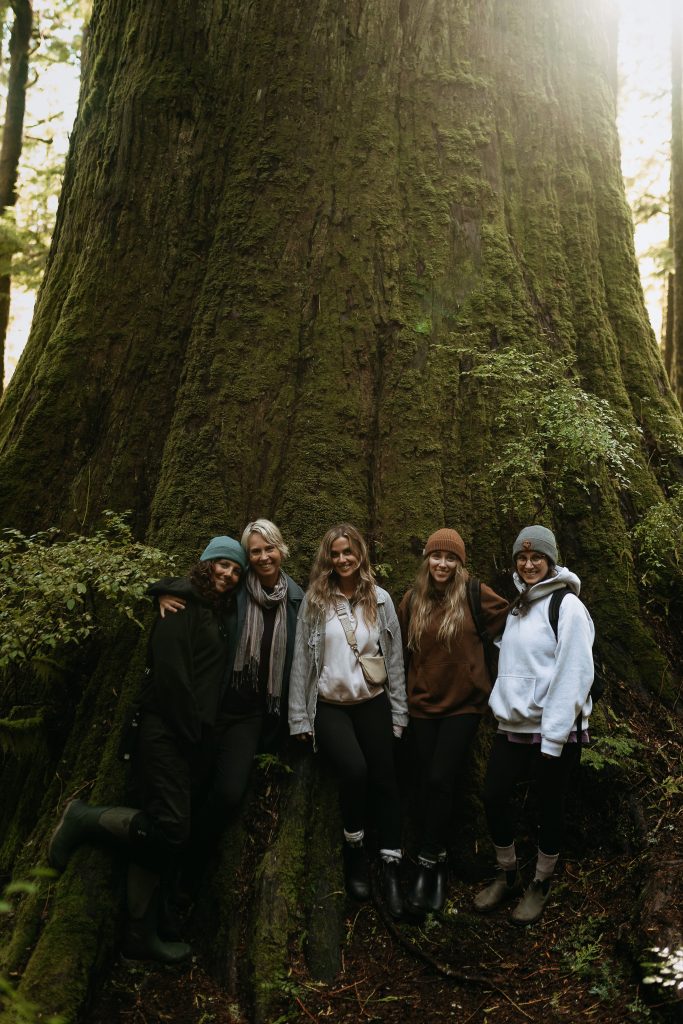 Five women stand smiling at the base of a giant moss-covered tree in a forest, dressed in outdoor clothing and boots, with sunlight filtering through the trees above.