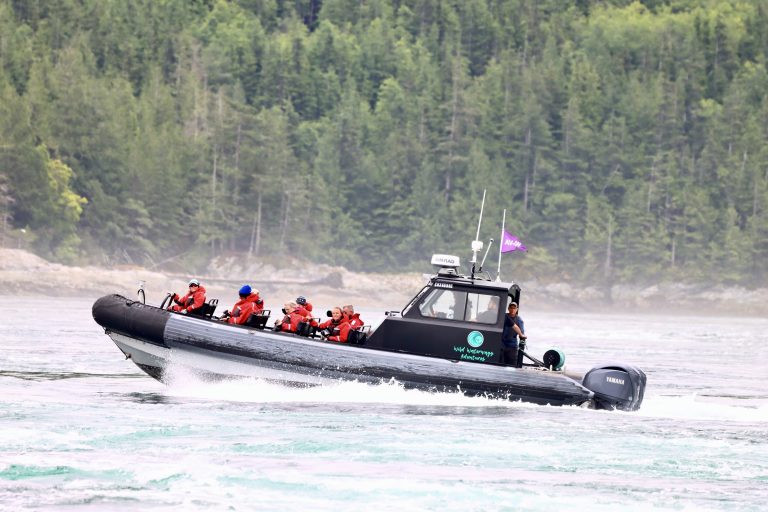 A black inflatable boat with several people in red life jackets speeds across the water, creating waves. Dense green forest and rocky shoreline are visible in the background.