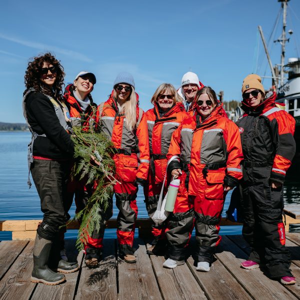 A group of seven people in bright orange and gray suits stand smiling on a wooden dock by the water, with boats behind them. One person holds a bundle of greenery, and the group appears ready for an outdoor adventure.