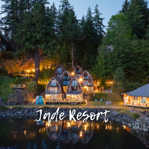 A lakeside resort with A-frame cabins and a round building, lit warmly at dusk, surrounded by tall trees; "Jade Resort" is written across the image.