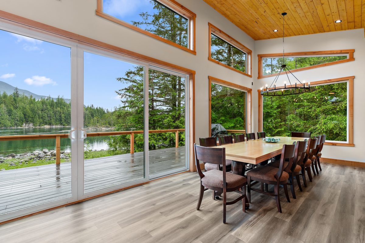 A modern dining room with large windows and sliding glass doors overlooking a scenic lake and forest, featuring a wooden table, chairs, and a chandelier under a wood-paneled ceiling.