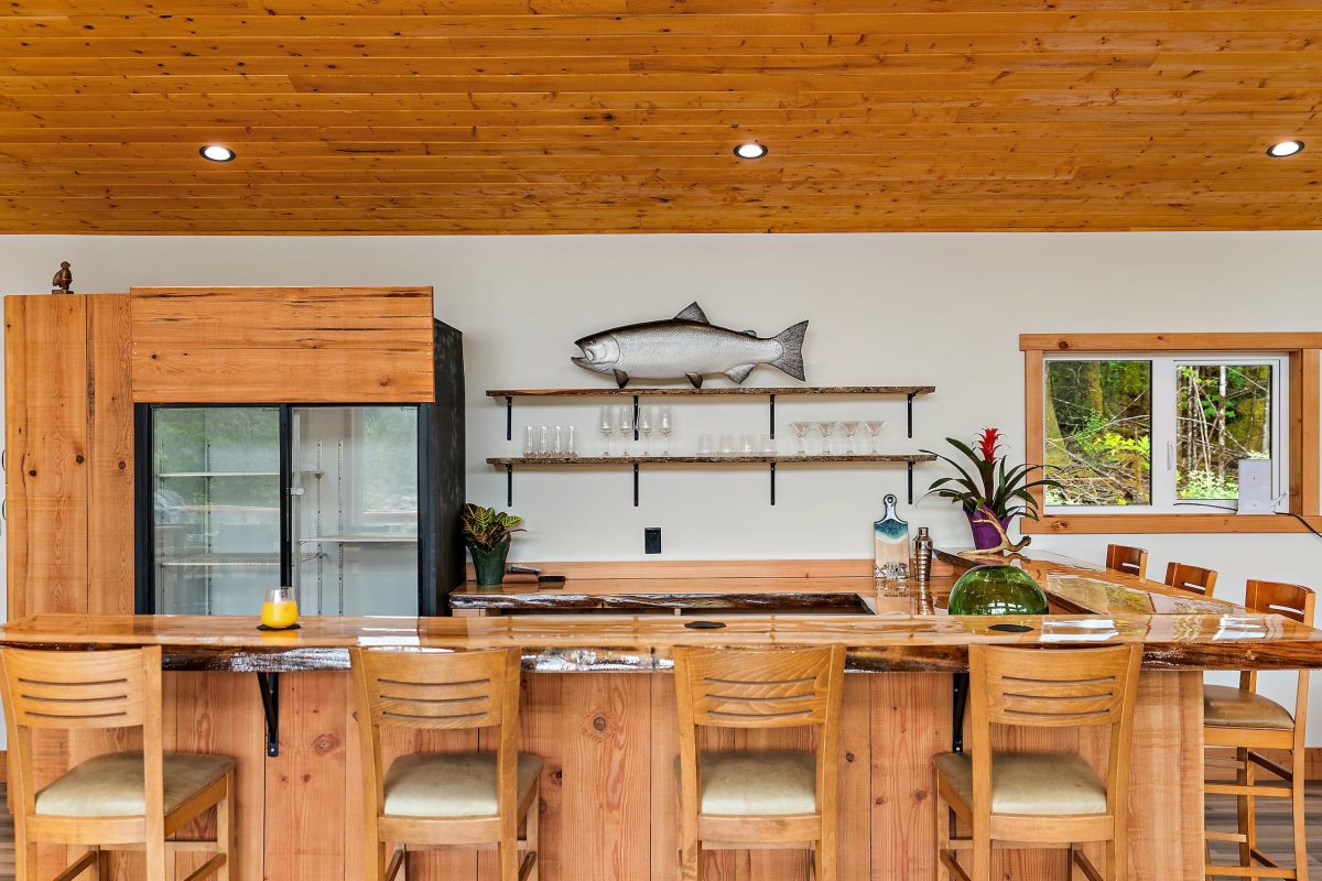 A rustic wooden bar with four chairs, a countertop with a drink and plants, glass shelves with glasses, a fish wall decoration, large fridge, and a window showing greenery outside.