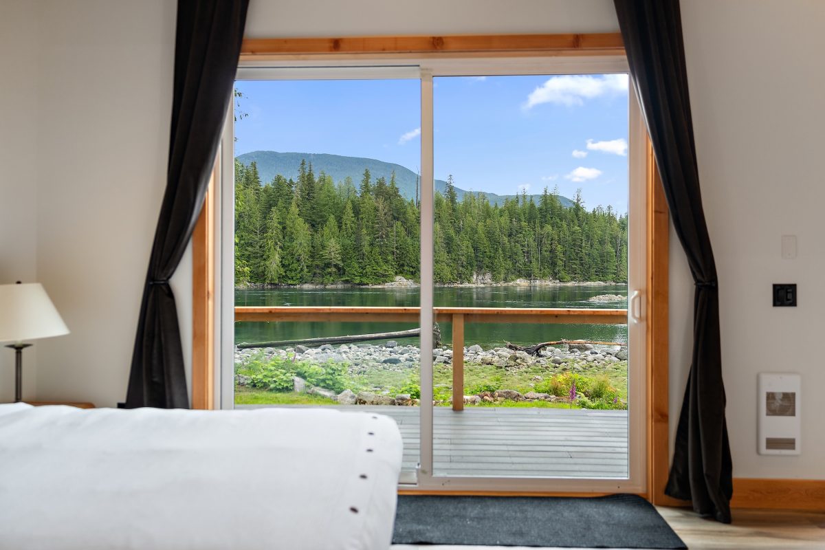 A bed near a sliding glass door opens to a deck with a view of a calm lake, green forest, and mountains under a blue sky with scattered clouds.