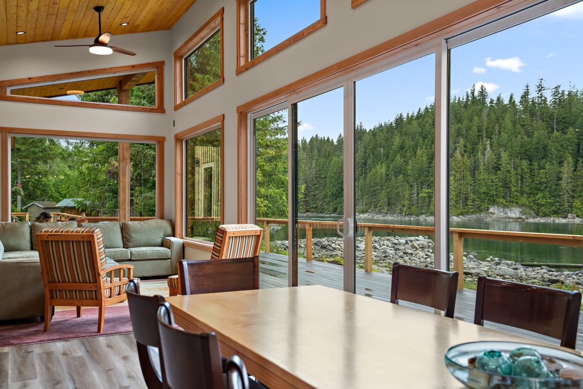 A modern living and dining area with wood accents, large windows, and sliding glass doors that overlook a scenic lake and forested hills, letting in abundant natural light.