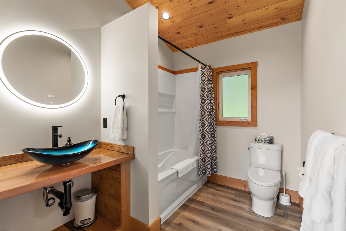 Modern bathroom with a round backlit mirror, vessel sink on a wooden counter, toilet, bathtub with a geometric shower curtain, wood-paneled ceiling, and a window above the toilet. Towels and a tissue box are visible.
