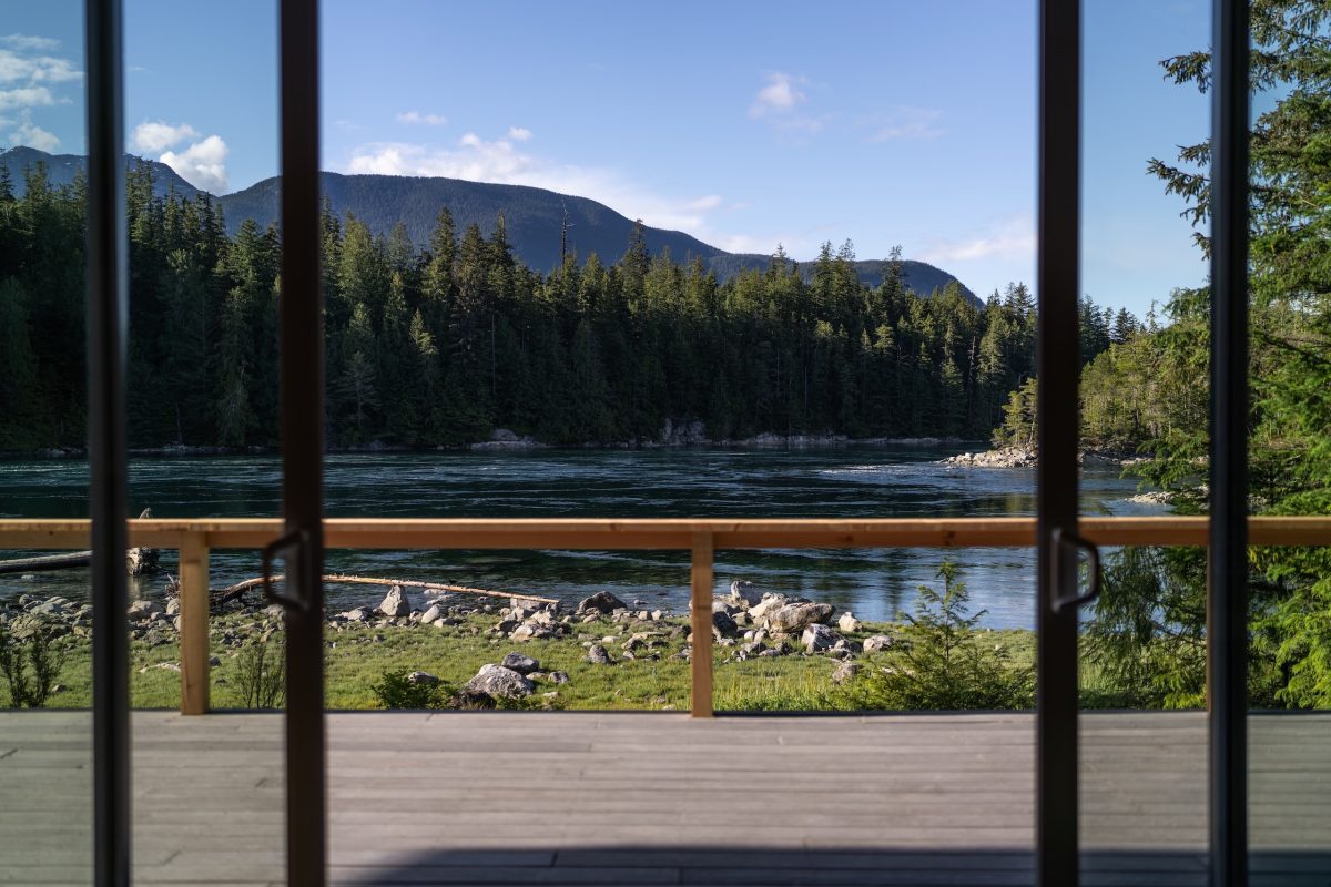 View through large glass doors showing a wooden deck, a calm river, forested area, and distant mountains under a blue sky with scattered clouds.