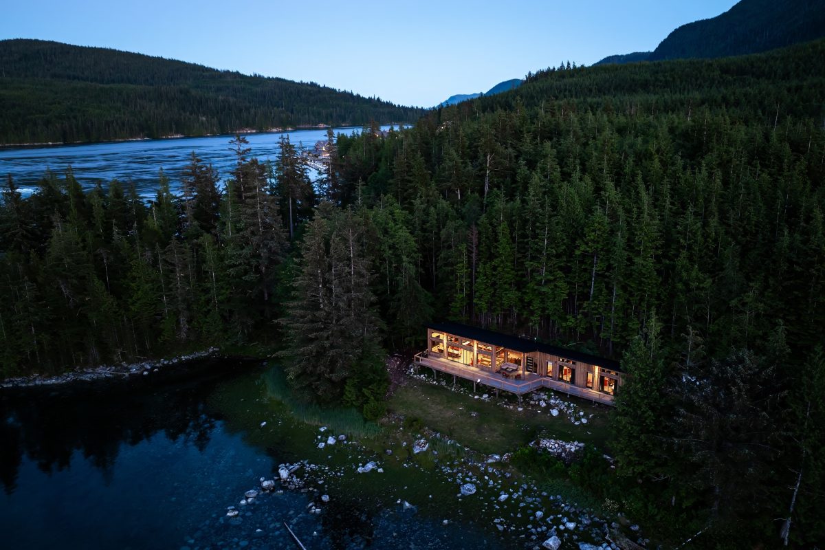 A modern cabin with large windows is nestled among dense evergreen trees by a clear river, with forested mountains in the background at dusk. Warm lights from the cabin stand out against the natural surroundings.