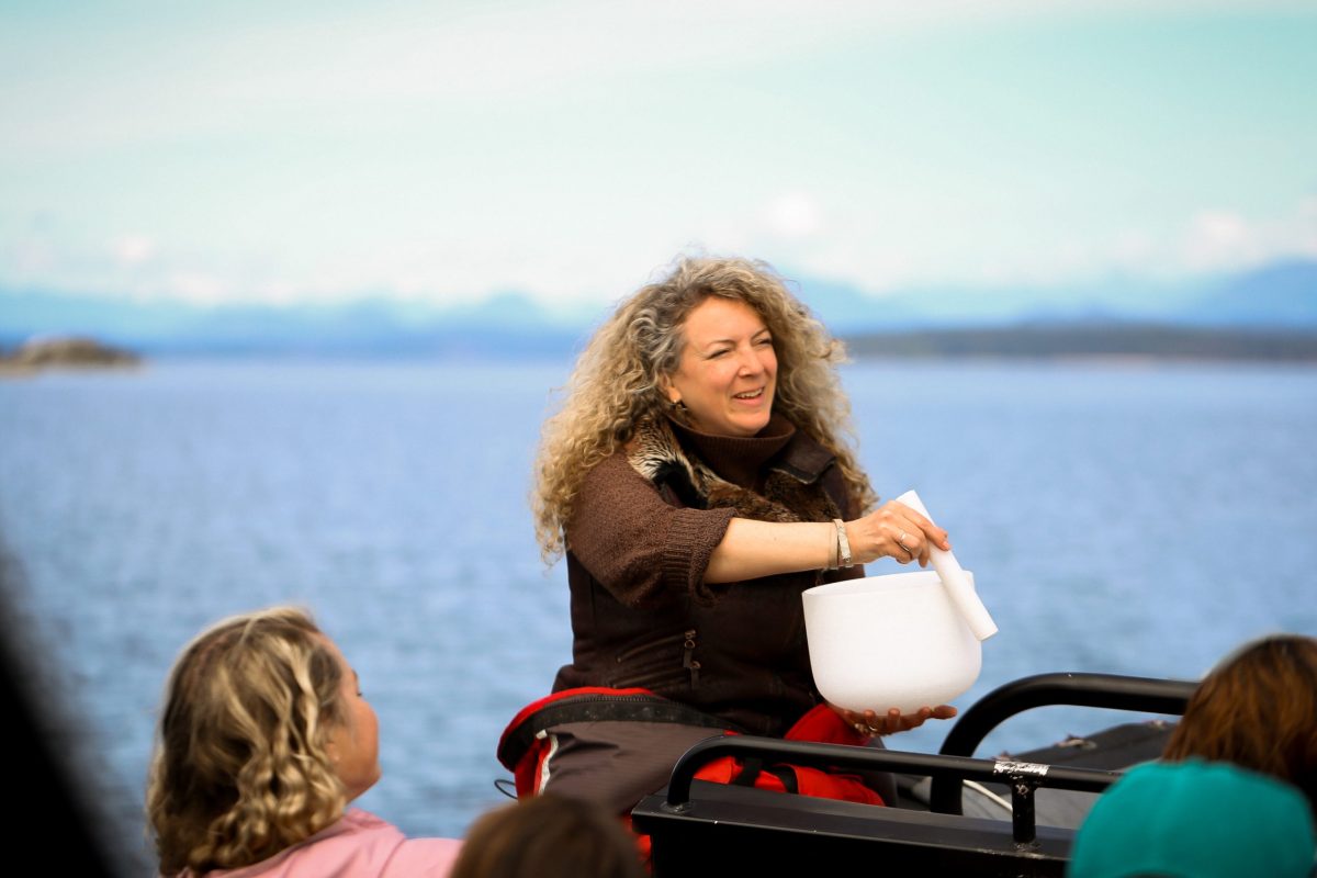A woman with curly blonde hair plays a white singing bowl outdoors by a body of water, with mountains in the background and two people watching her.