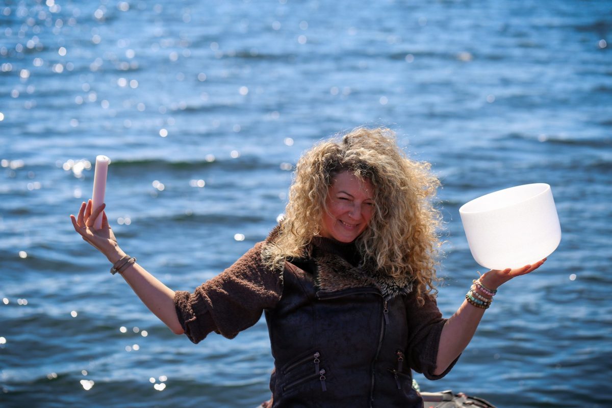 A woman with curly hair stands by a body of water, smiling. She holds a white crystal singing bowl in one hand and a stick in the other, with sunlight reflecting off the water in the background.