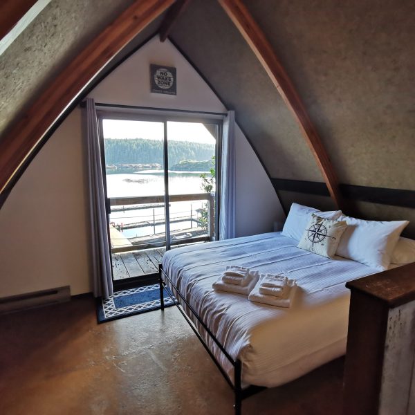 A cozy A-frame bedroom with a neatly made bed, fresh towels, and a decorative pillow, facing a glass door that opens to a balcony with a scenic view of water and forested hills.