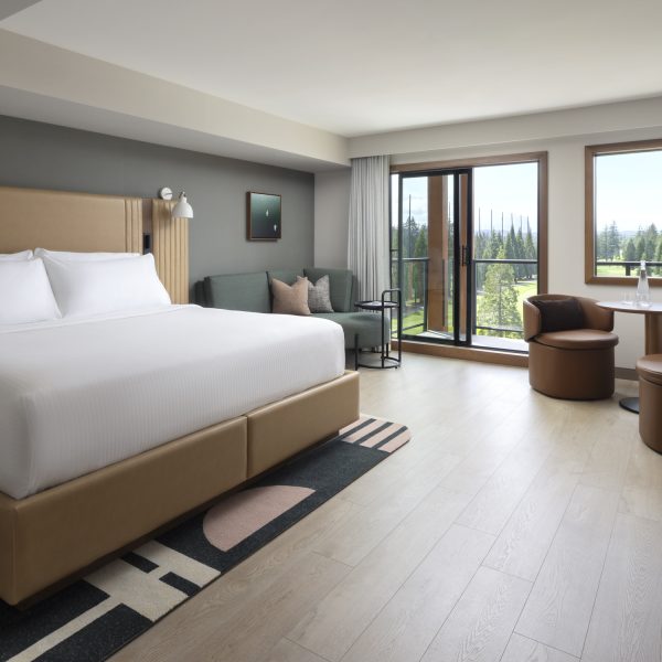 Modern hotel room with a large bed, sofa, small round table with two chairs, TV, potted plant, artwork, and a large window with a balcony overlooking greenery. The decor is neutral with wood and leather accents.