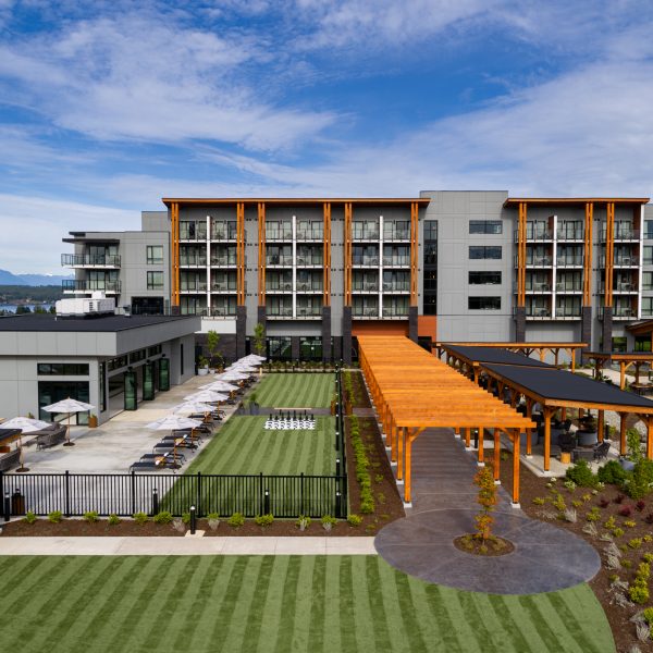 A modern hotel with large windows and balconies, features a wooden pergola walkway, outdoor seating area with umbrellas, landscaped lawns, and views of distant mountains under a blue sky.