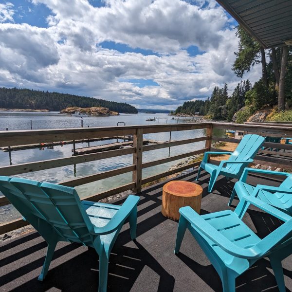 Four turquoise Adirondack chairs and a round wooden table sit on a deck overlooking a calm bay with forests and clouds in the background, creating a peaceful, scenic waterfront view.