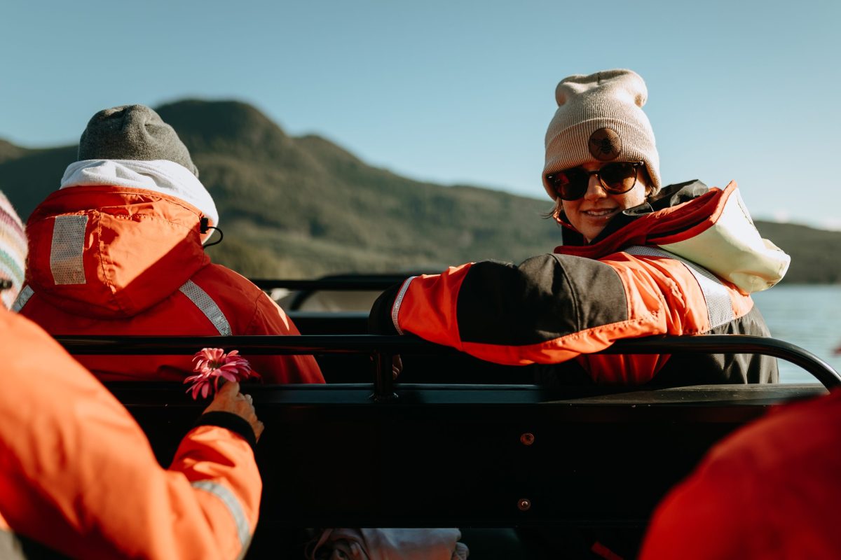 A person in an orange jacket and beanie smiles while sitting on a boat, looking back over their shoulder. Another person holds a pink flower. Mountains and water are visible in the background under a clear sky.