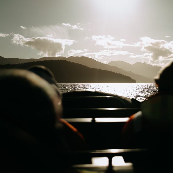 Two people sit in a boat on a lake, facing distant mountains under a bright sun. The water reflects the sunlight and the landscape is silhouetted, creating a peaceful and scenic atmosphere.