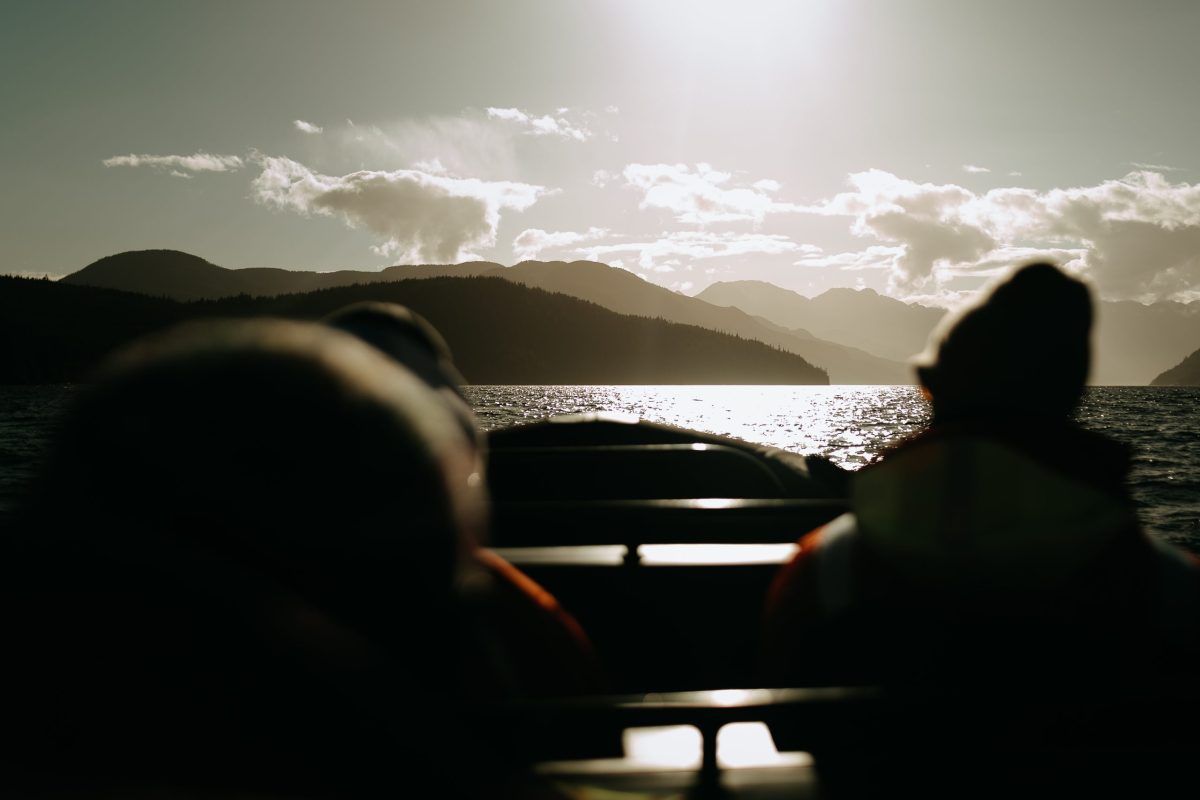 Two people sit in a boat on a lake, facing distant mountains under a bright sun. The water reflects the sunlight and the landscape is silhouetted, creating a peaceful and scenic atmosphere.