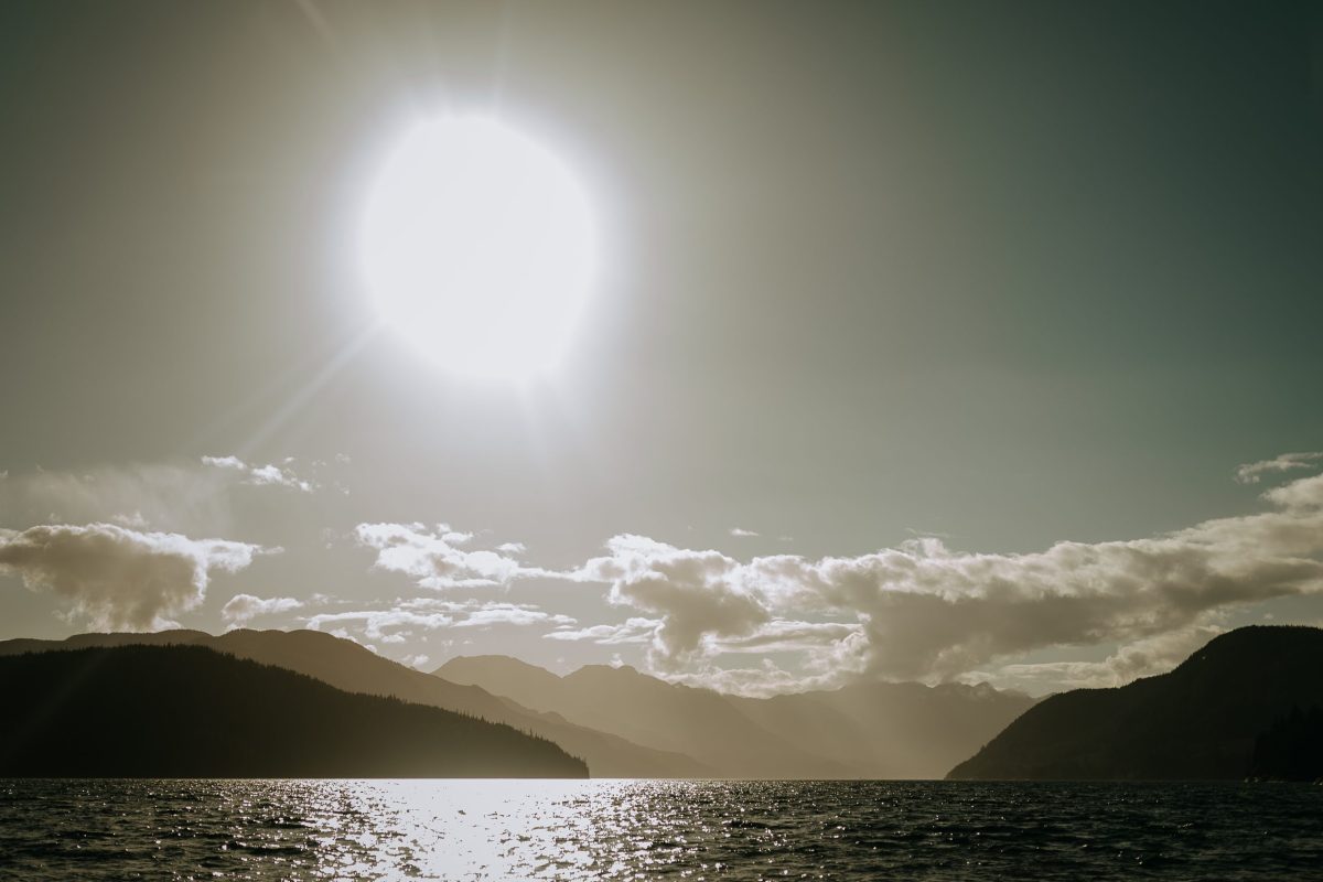 The sun shines brightly over a serene lake, with its light reflecting on the water. Mountains and clouds are visible in the distance, creating a peaceful and scenic landscape.