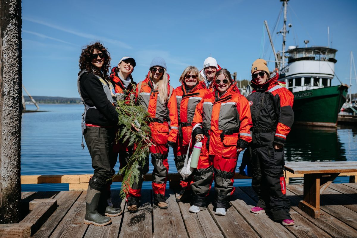 A group of seven people wearing orange and black insulated suits stand on a wooden dock by the water, smiling at the camera; boats and blue sky are visible in the background.
