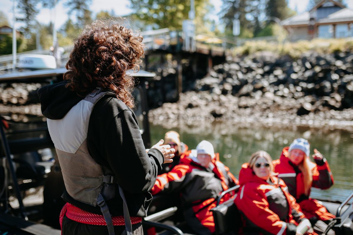 A person with curly hair and a life jacket faces a group of four people in red waterproof suits seated in a boat on the water, near a rocky shore with trees and buildings in the background.