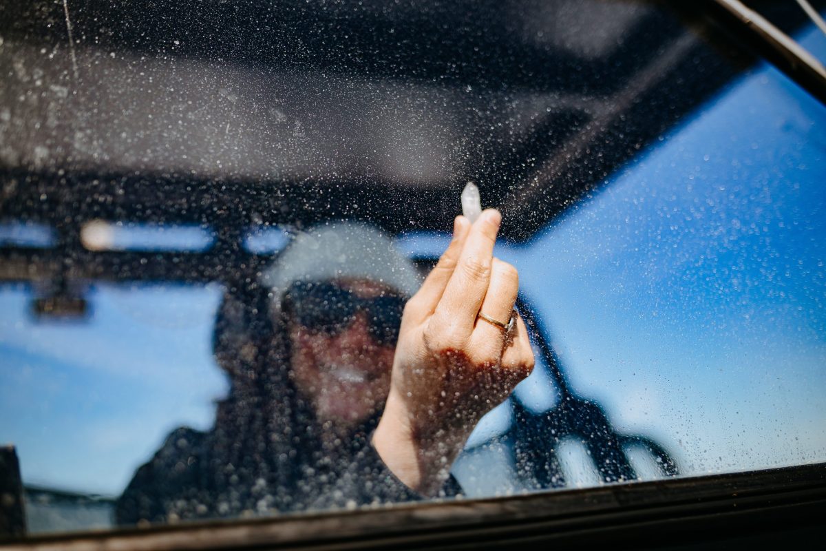 A person in sunglasses and a beanie wipes a dirty, water-spotted windshield with a small cloth, captured from the other side of the glass on a bright, clear day.