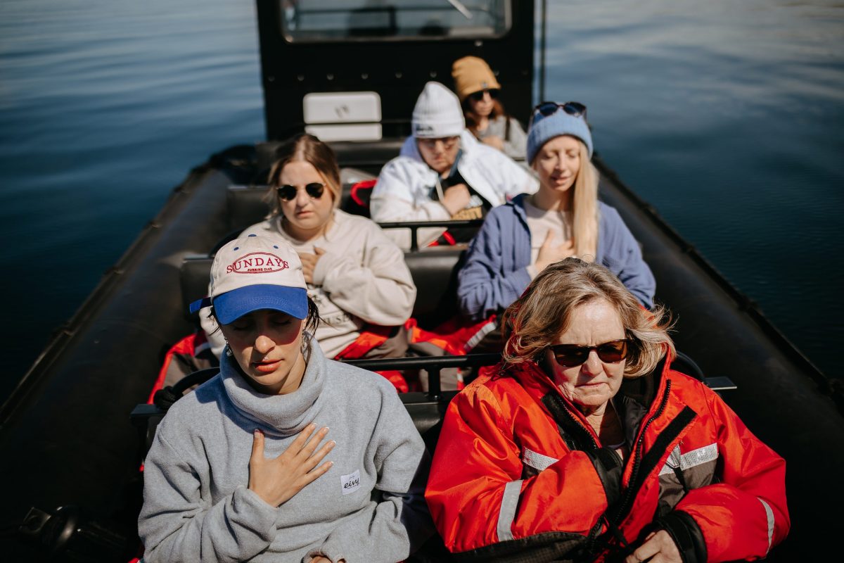 Six people sit in rows on a boat, eyes closed and hands on their chests, appearing calm and reflective. The water around them is still, and they wear casual, warm clothing including jackets, hats, and sunglasses.