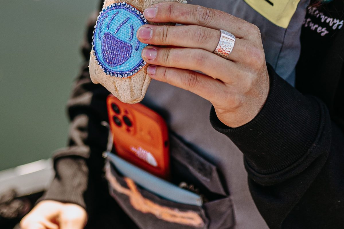 A person wearing a gray vest holds a small pouch with a blue beaded design. An orange phone is visible in the vest pocket. The background appears to be outdoors, possibly near water.