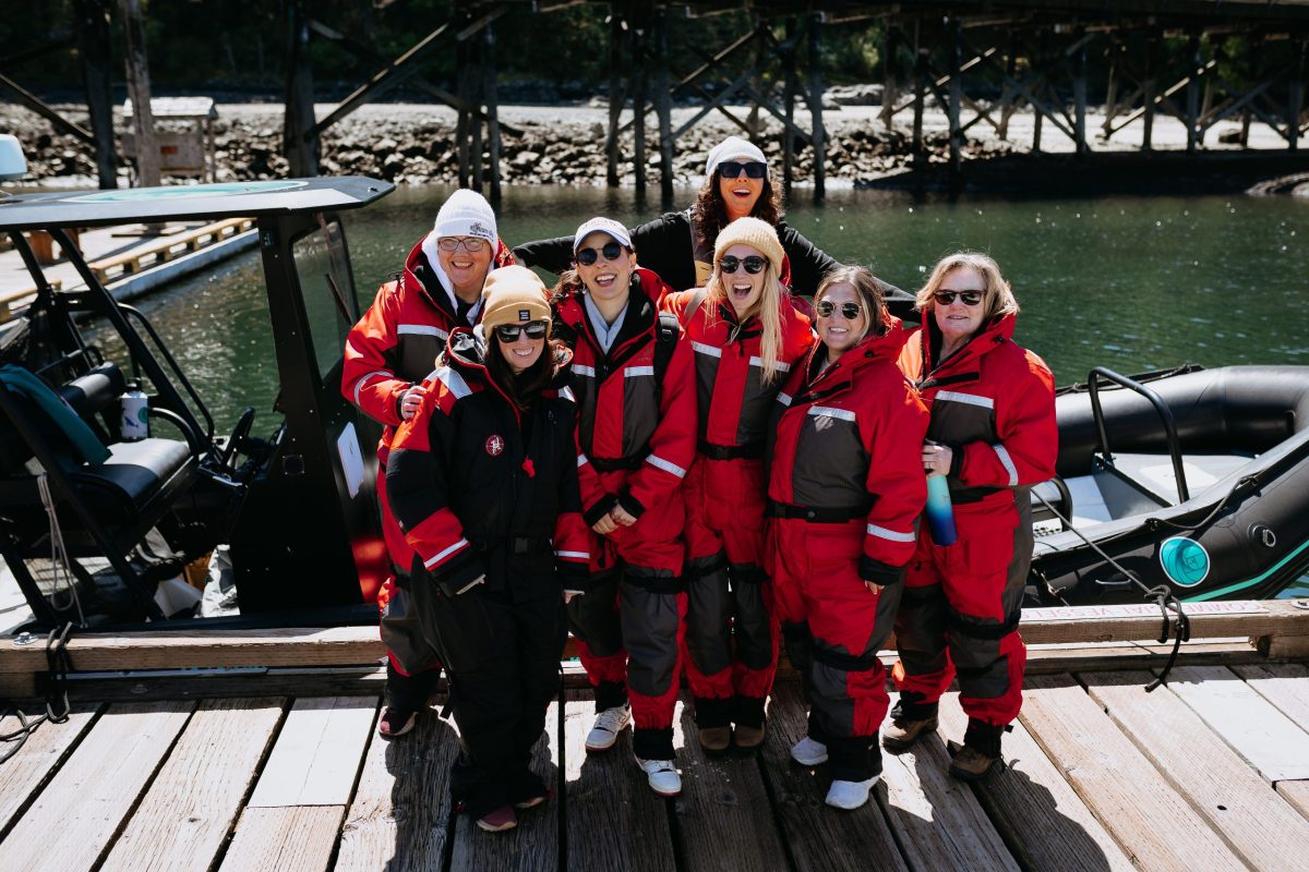 A group of seven people wearing red and black cold-weather suits stand on a wooden dock in front of an inflatable boat, smiling at the camera with water and trees in the background.