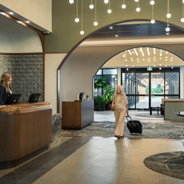 A woman with a suitcase walks into a modern hotel lobby toward the reception desk, where a receptionist stands ready to assist. The lobby is well-lit with hanging lights and stylish decor.