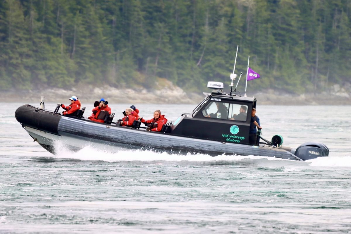 A black inflatable boat with six people in orange life jackets speeds across the water, surrounded by forested shoreline. Some passengers are taking photos, and a purple flag is flying on the boat.