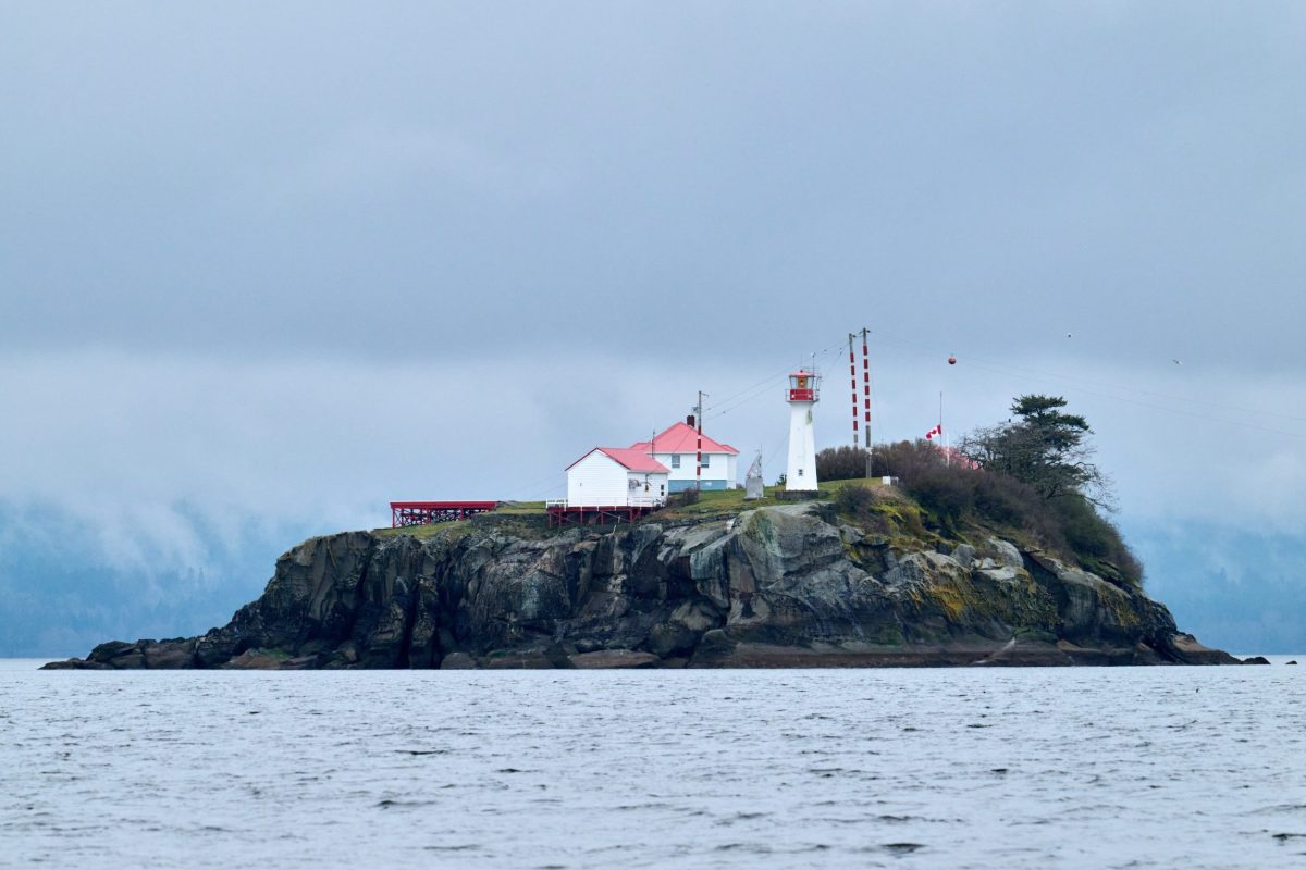 A small rocky island with a white lighthouse and buildings with red roofs, surrounded by calm water and cloudy skies, with trees and vegetation on the island.